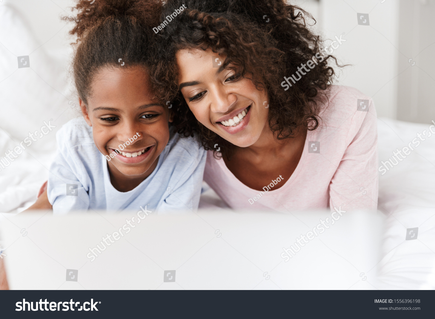 Smiling young mother and her little daughter wearing pajamas releaxing on bed, using laptop computer, chatting #1556396198