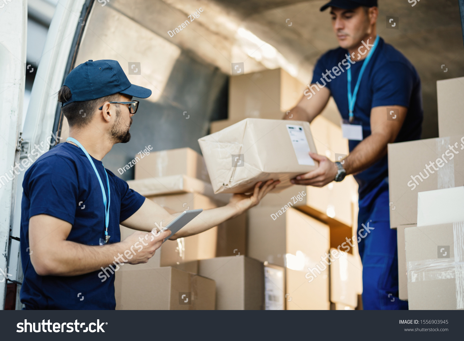 Young courier using touchpad while loading packages with his coworker in a delivery van.  #1556903945