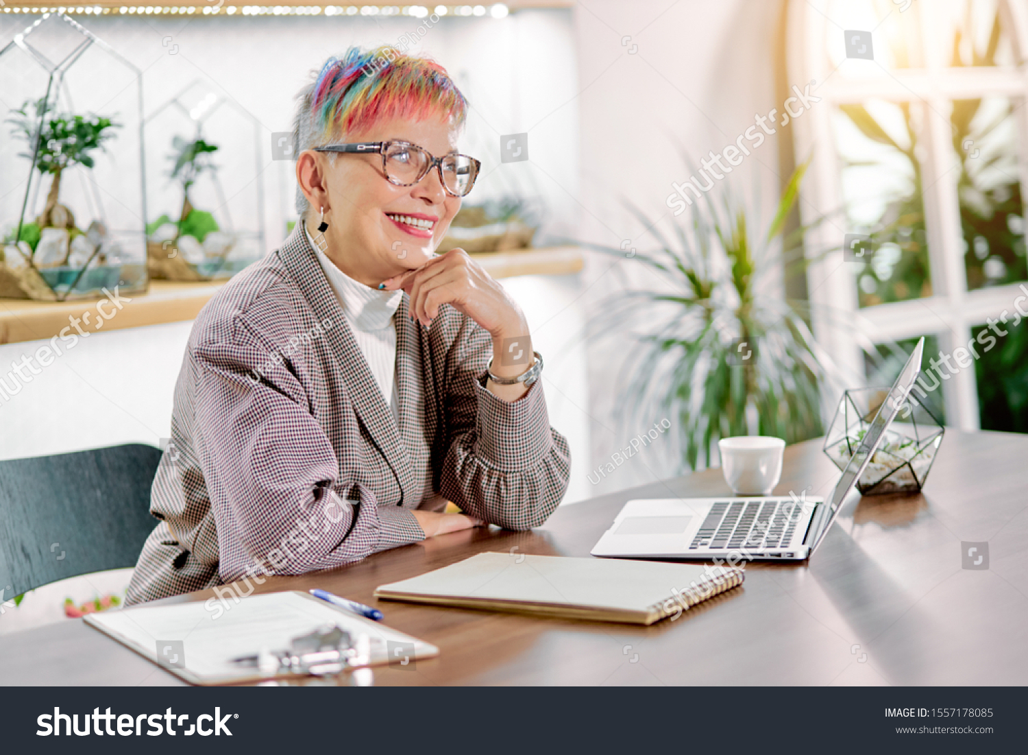 Portrait of well-favoured woman involved in business work smile, wearing blazer, glasses, short colorful hair. Isolated in light office, plants background #1557178085