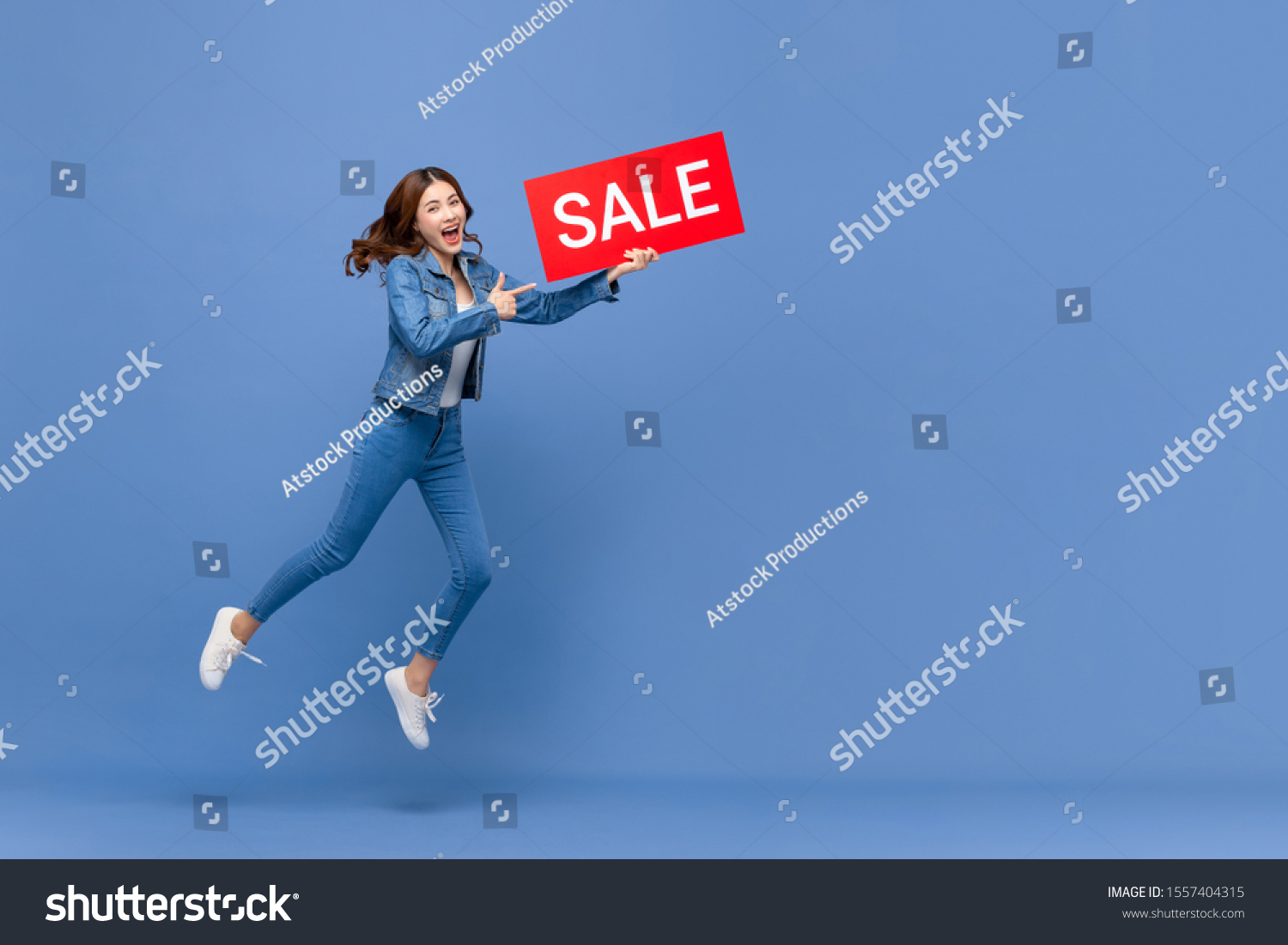 Excited Asian woman in casual jean clothes jumping with red sale sign in hand isolated on light blue background with copy space #1557404315