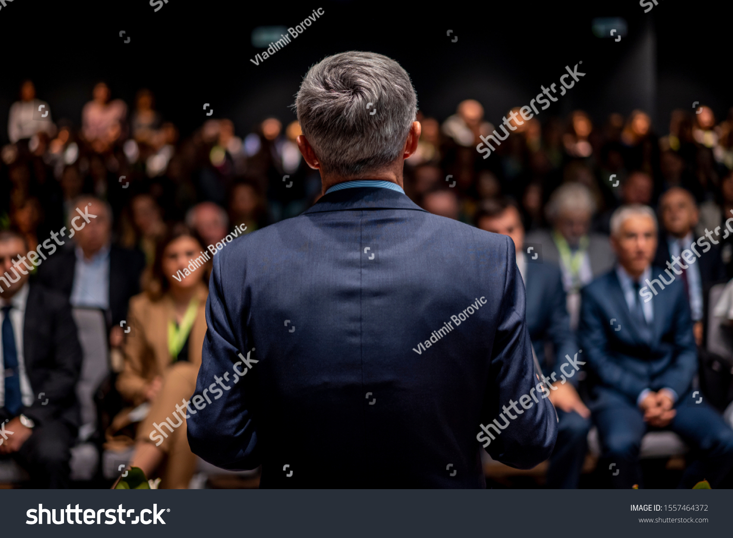 Business man is making a speech in front of a big audience at a conference hall. Speaker giving a talk on corporate business or political conference. Politician talking to group of people #1557464372
