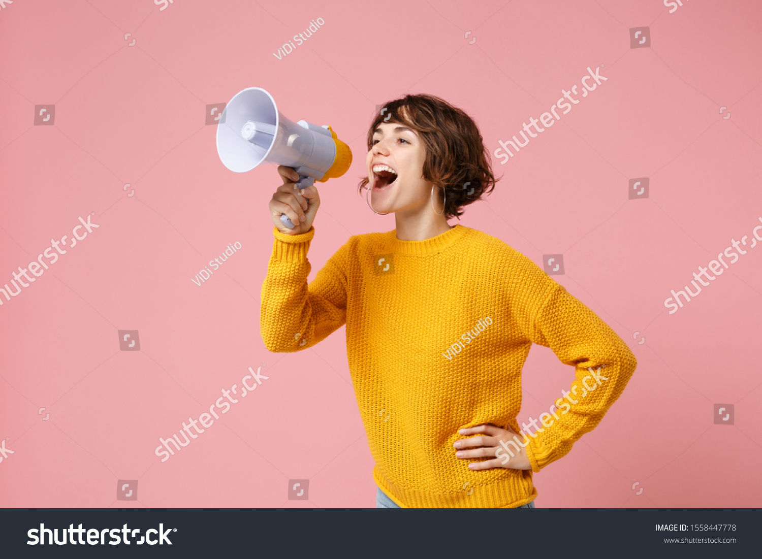 Funny young brunette woman girl in yellow sweater posing isolated on pastel pink wall background studio portrait. People sincere emotions lifestyle concept. Mock up copy space. Screaming in megaphone #1558447778