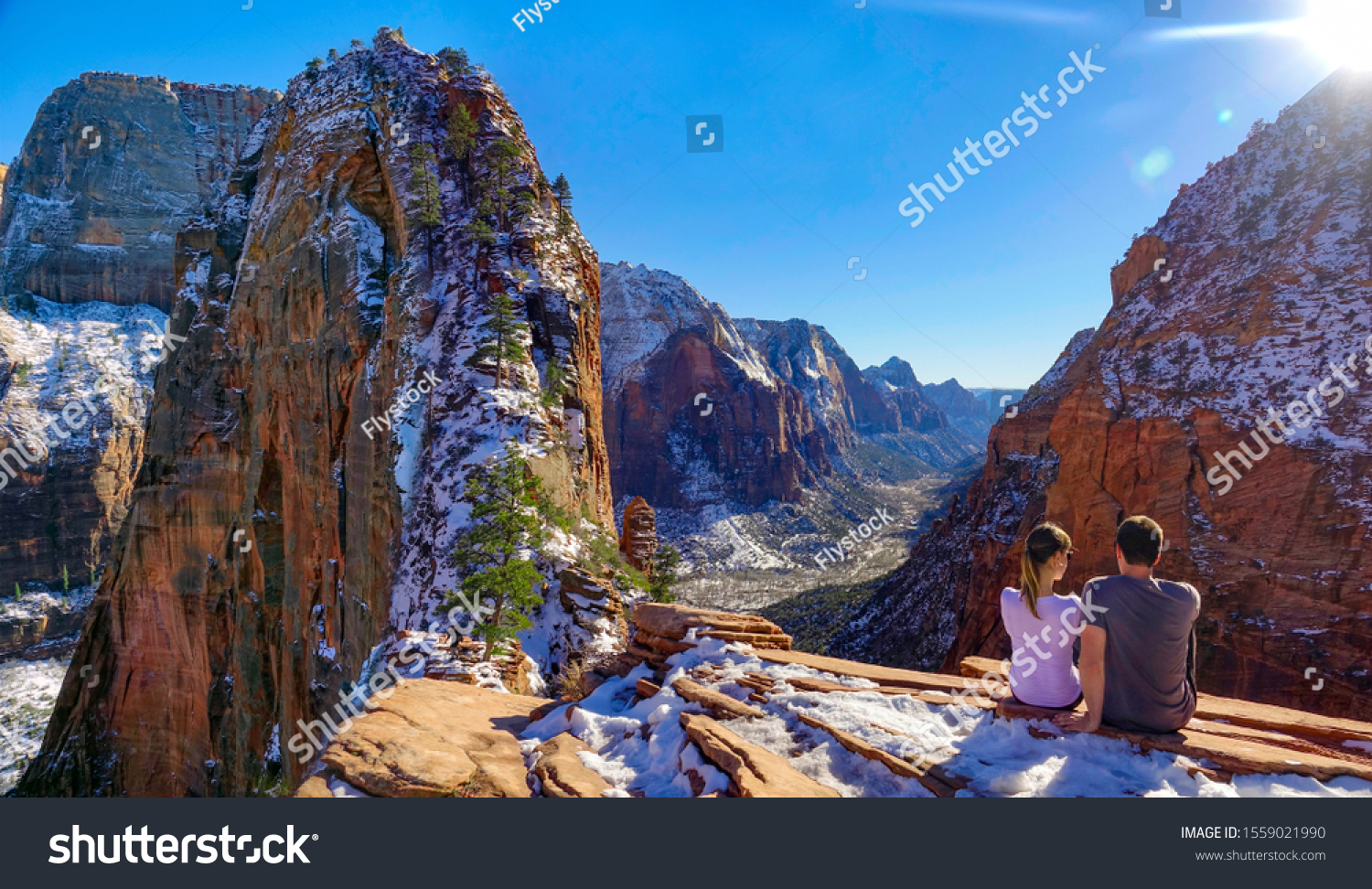 COPY SPACE, LENS FLARE: Young couple sits atop Angel's Landing and observes the breathtaking canyon in Utah. Tourist observe the valley and red sandstone mountains after a successful hike in Zion Park #1559021990