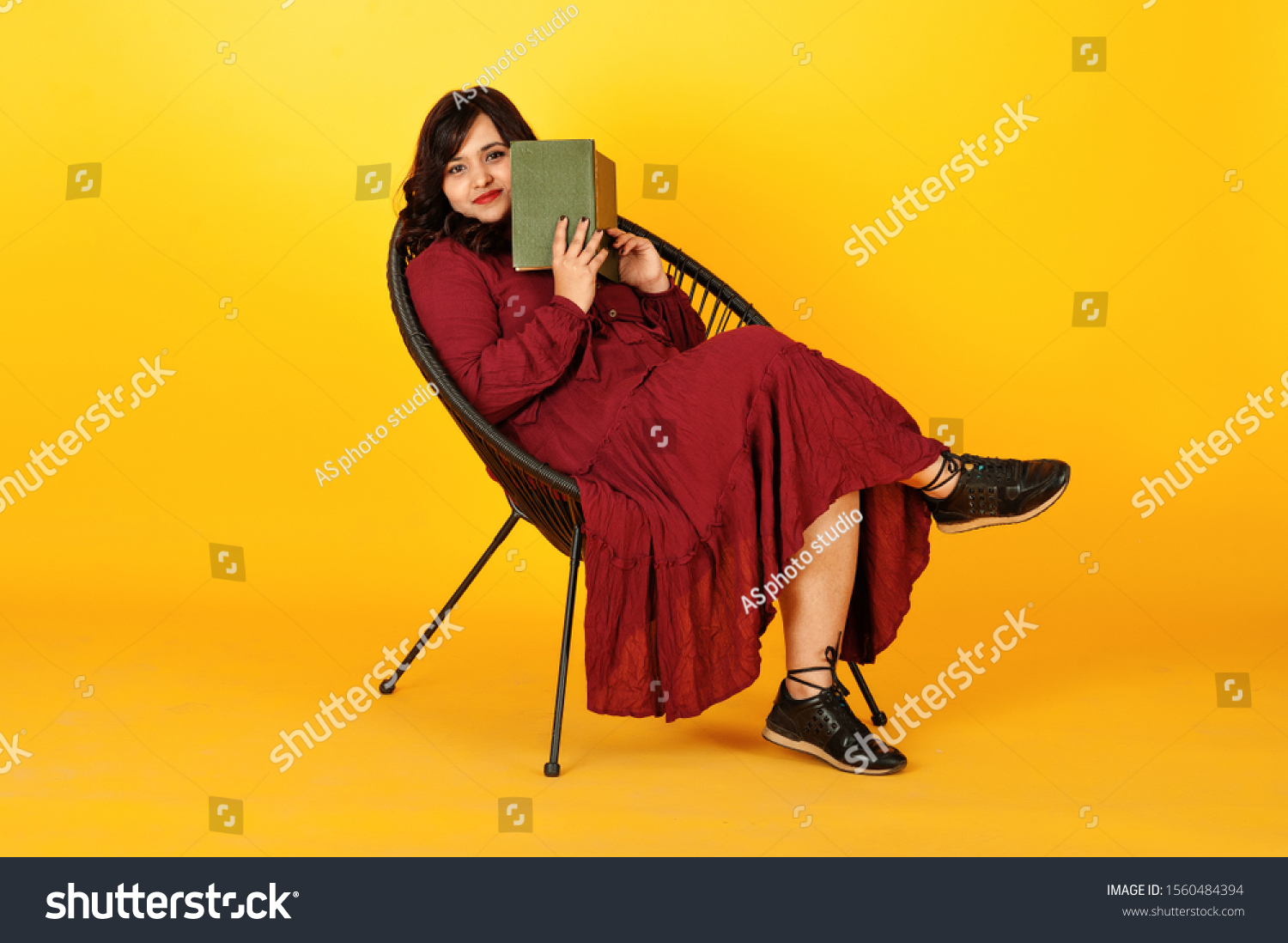 Attractive south asian woman in deep red gown dress posed at studio on yellow background sitting on chair with book. #1560484394