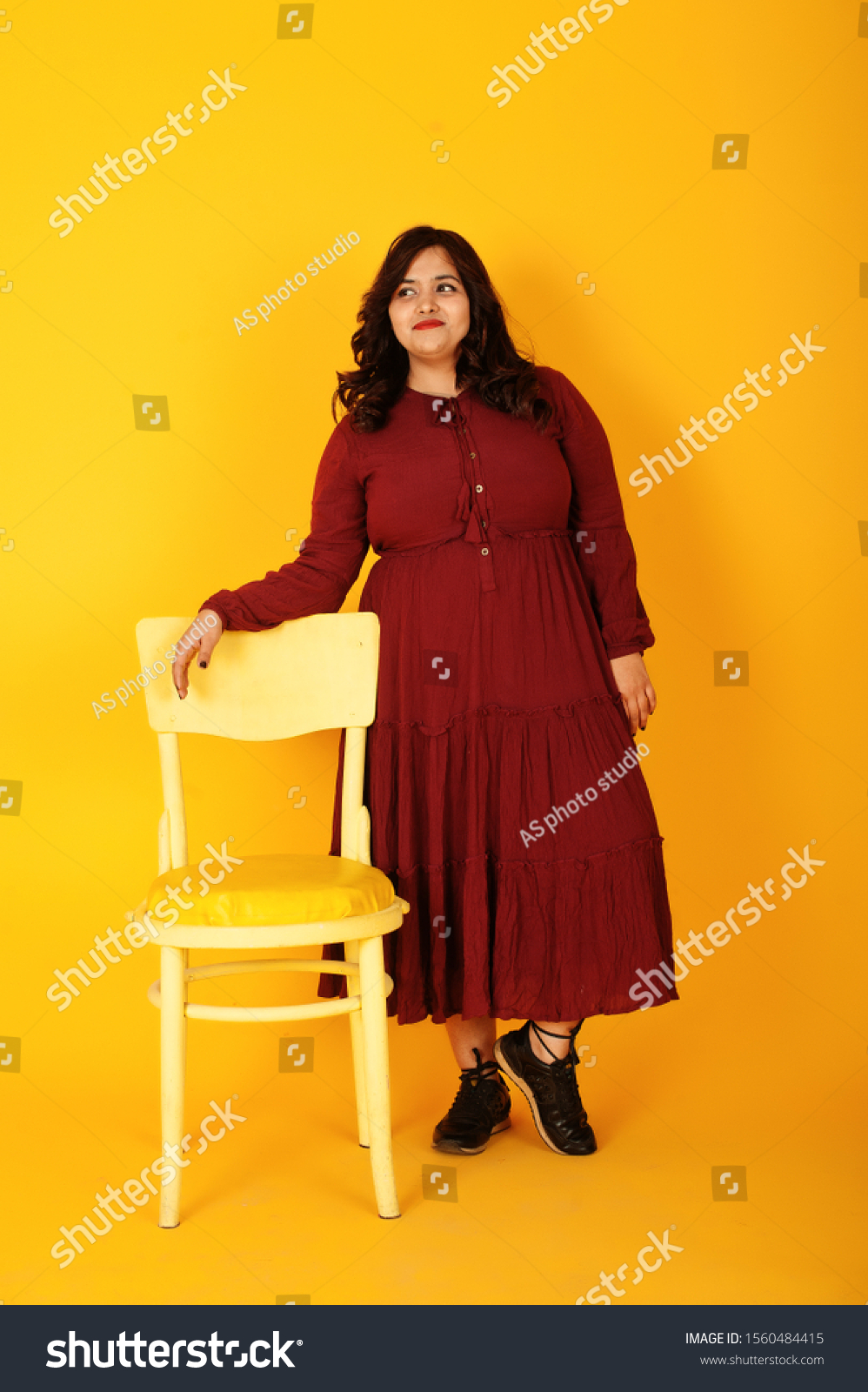 Attractive south asian woman in deep red gown dress posed at studio on yellow background with chair. #1560484415