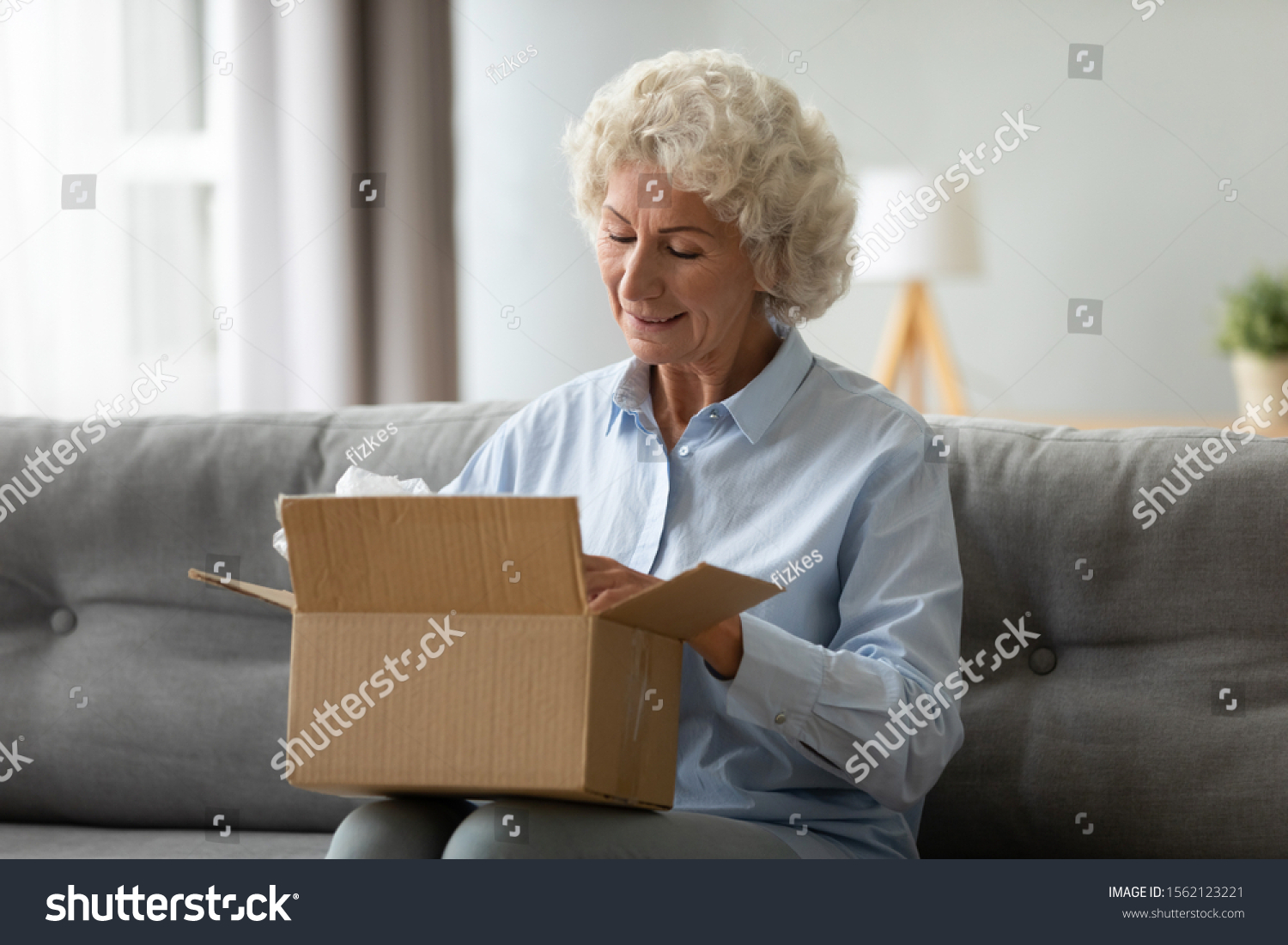 Smiling elderly woman customer receive post shipment parcel at home, happy old senior grandma hold open cardboard box sit on sofa in living room, online shopping order fast courier delivery concept #1562123221