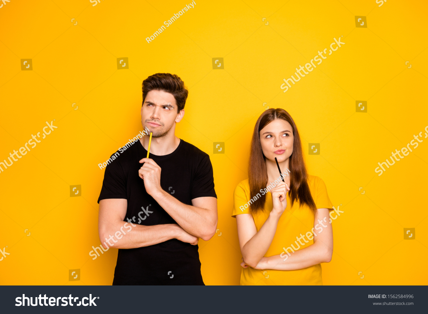 Photo of serious confident couple of two people interested in thinking over standing under empty space holding pens wearing black t-shirt isolated over vivid yellow color background #1562584996