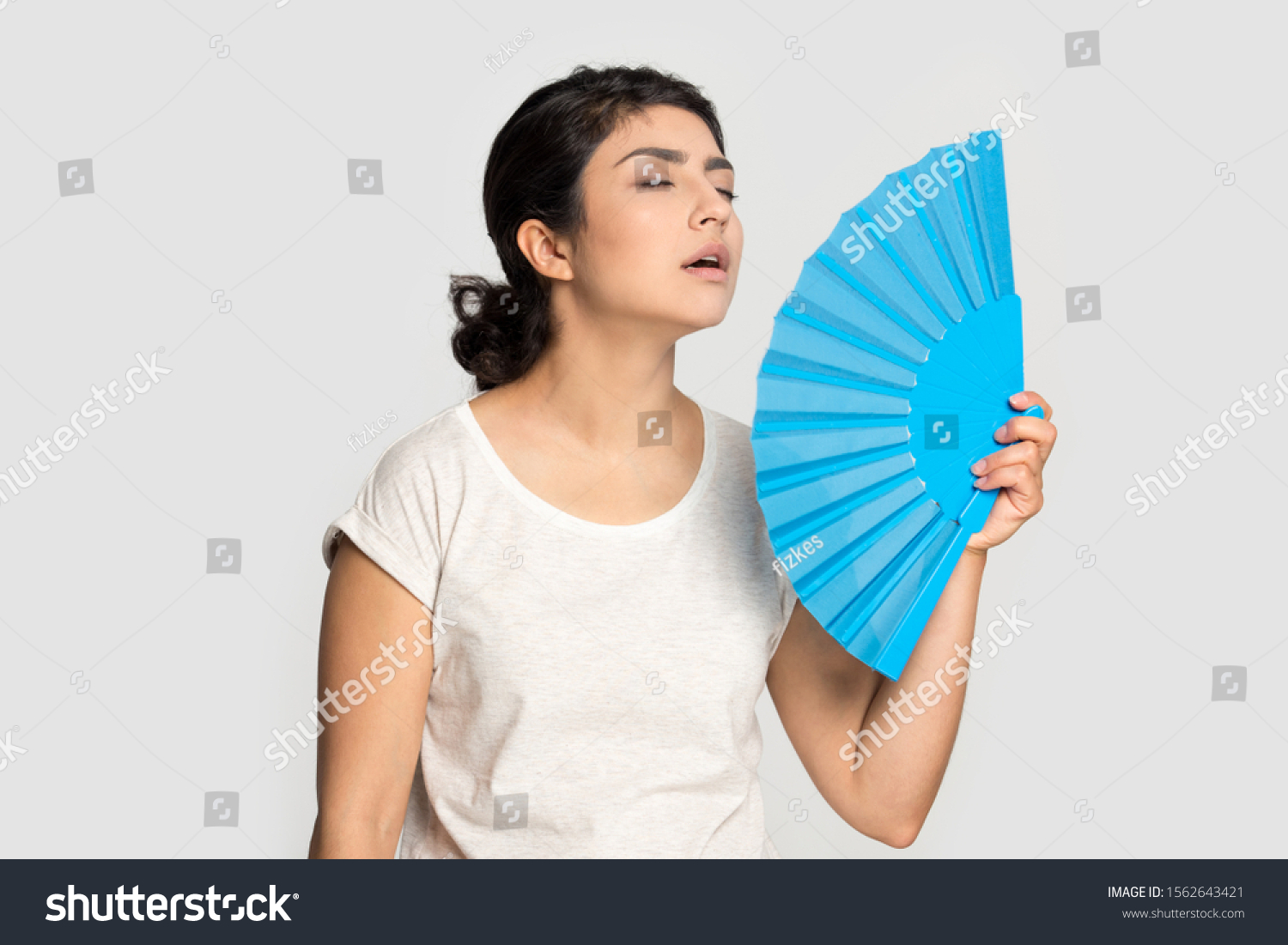 Tired exhausted Indian girl waving blue paper fan standing isolated on grey background, suffering from heat, sweaty young woman cooling in hot summer weather, high temperature, close up #1562643421