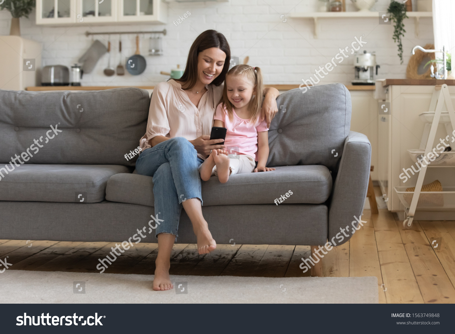 Full length front view happy 30s woman sitting on sofa with preschool daughter, looking at cellphone screen, watching funny cartoons, making selfie, holding call or recording video for social network. #1563749848