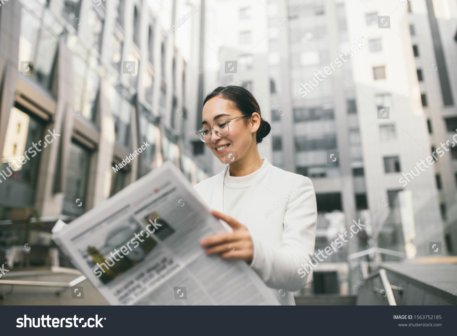 Young attractive Asian female banker or accountant in glasses is reading newspaper outside a modern office center or a bank #1563752185