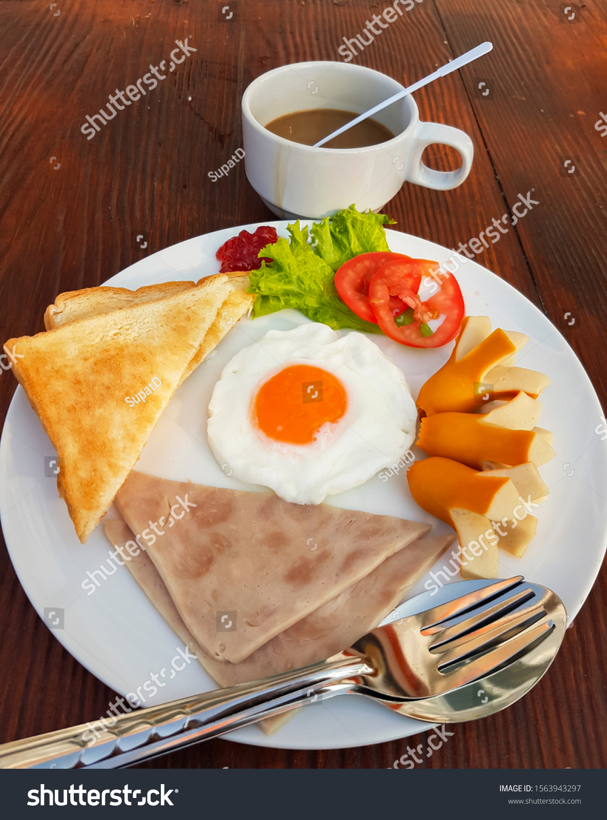 American breakfast includes fried egg, filling, ham, toast, lettuce and tomato, sliced on a white plate and coffee on a wooden table. #1563943297