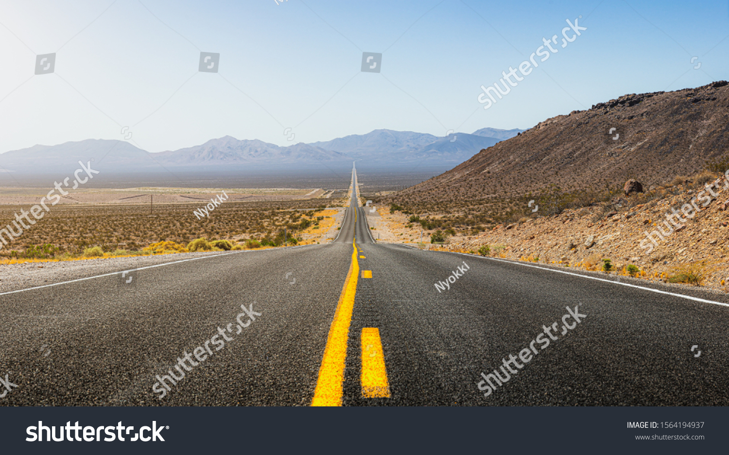 Beautiful panoramic view of a long straight road cutting through a barren scenery of the wild American Southwest with extreme heat haze on a hot and sunny day with blue sky in summer #1564194937