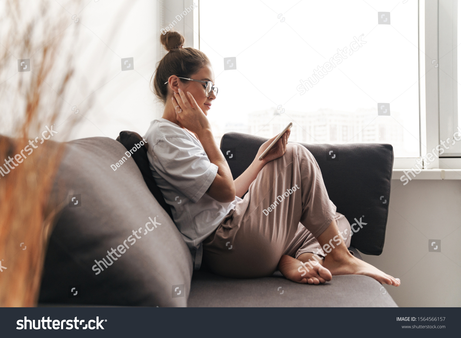 Image of concentrated nice woman in eyeglasses using cellphone while sitting on sofa at living room #1564566157