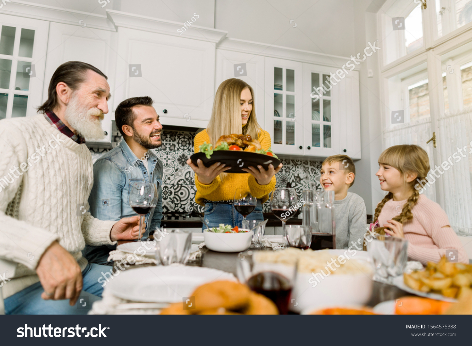 The family sits down for dinner on Thanksgiving. Young woman serves a festive turkey with a salad, grandfather, father and children sit and look at the tasty food and smile #1564575388