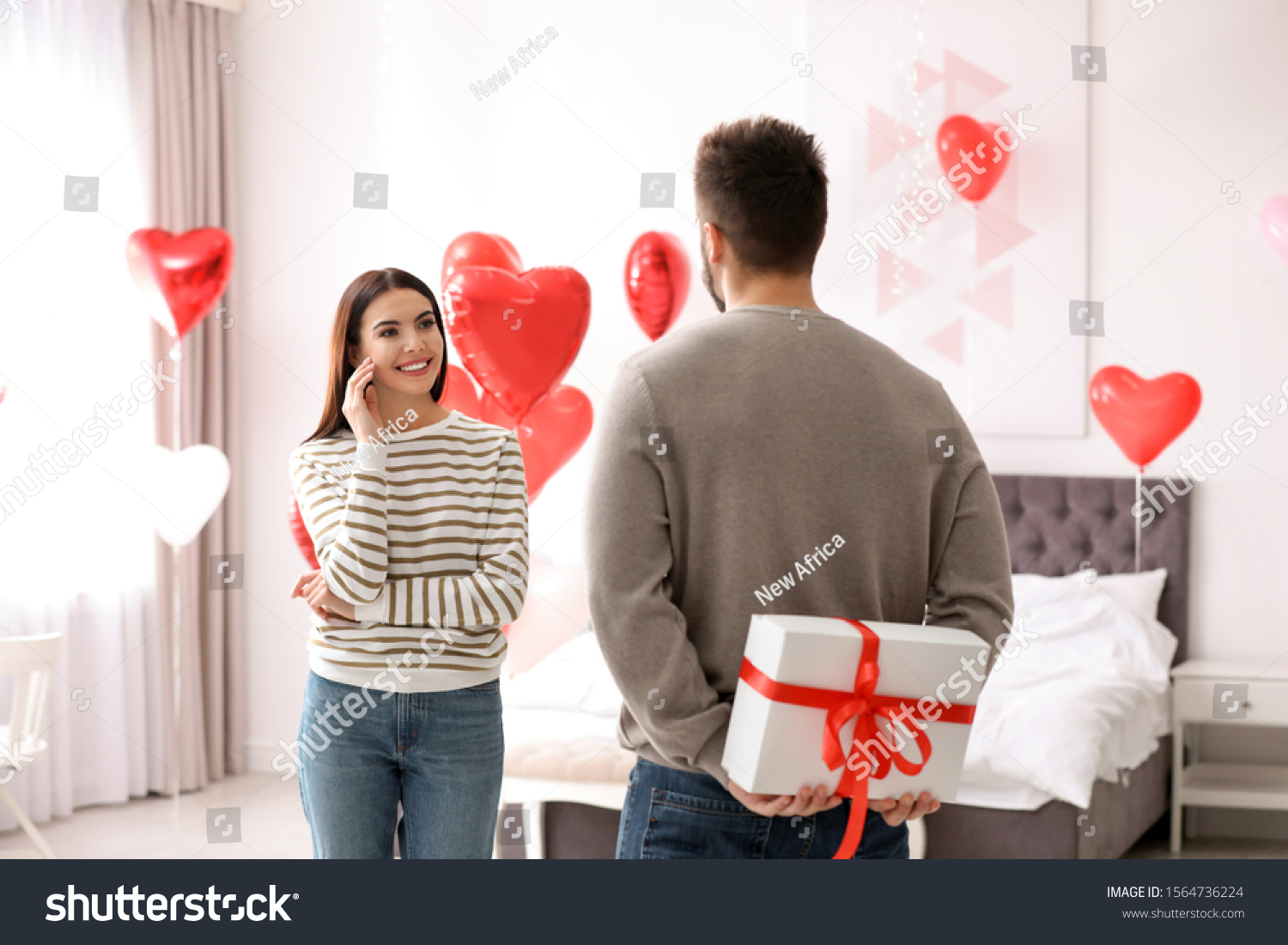 Young man presenting gift to his girlfriend in bedroom decorated with heart shaped balloons. Valentine's day celebration #1564736224