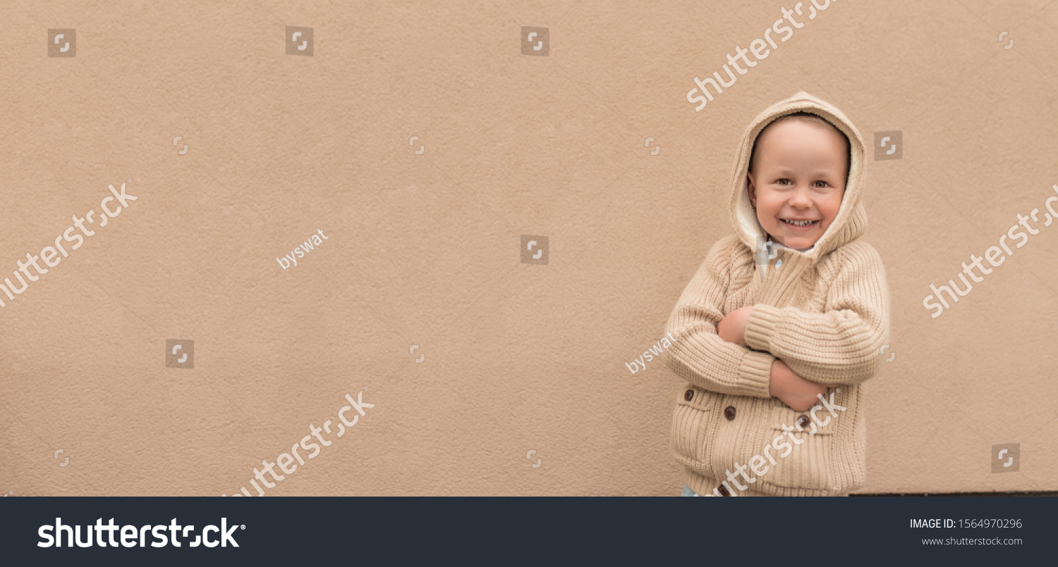Little boy 3-5 years old, beige sweater with hood, happy smiling, cheerful joyful. Free space for text. Background wall, autumn on street. Emotions pleasure fun. Holiday birthday concept #1564970296