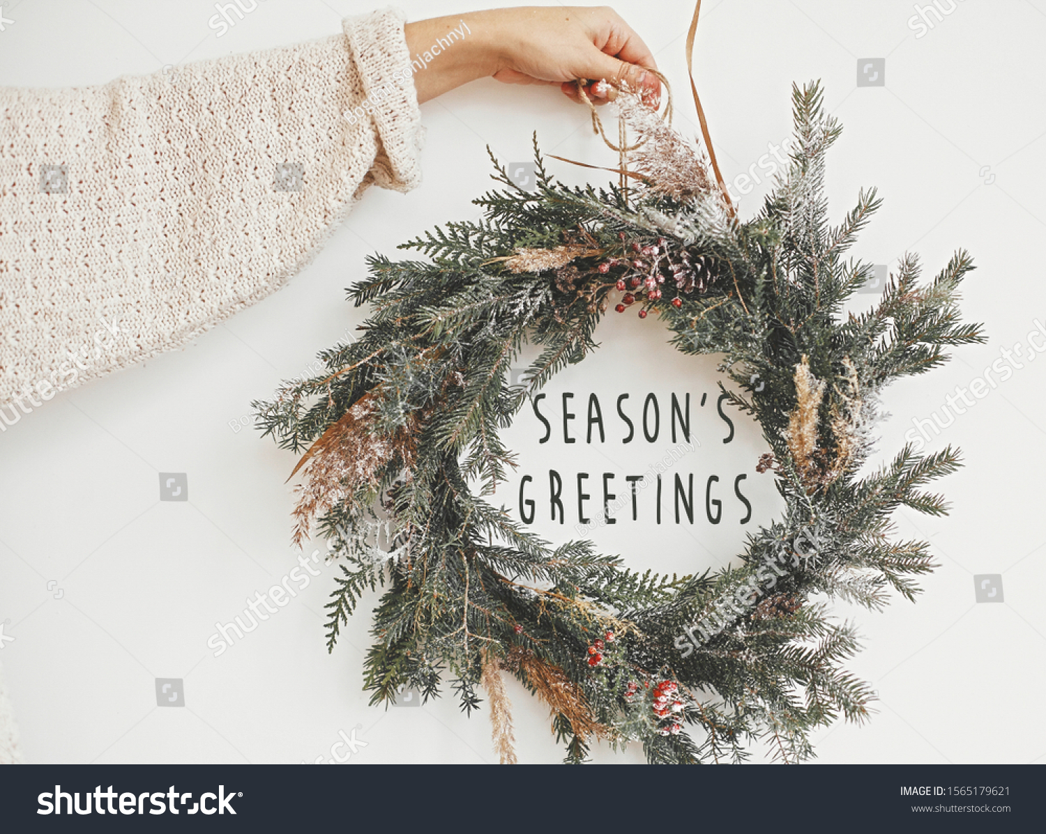 Season's greetings text sign on christmas rustic wreath in girl hand. Creative rural  wreath with fir branches, berries, pine cones, herbs hanging on white wall in room. Seasons greeting card #1565179621