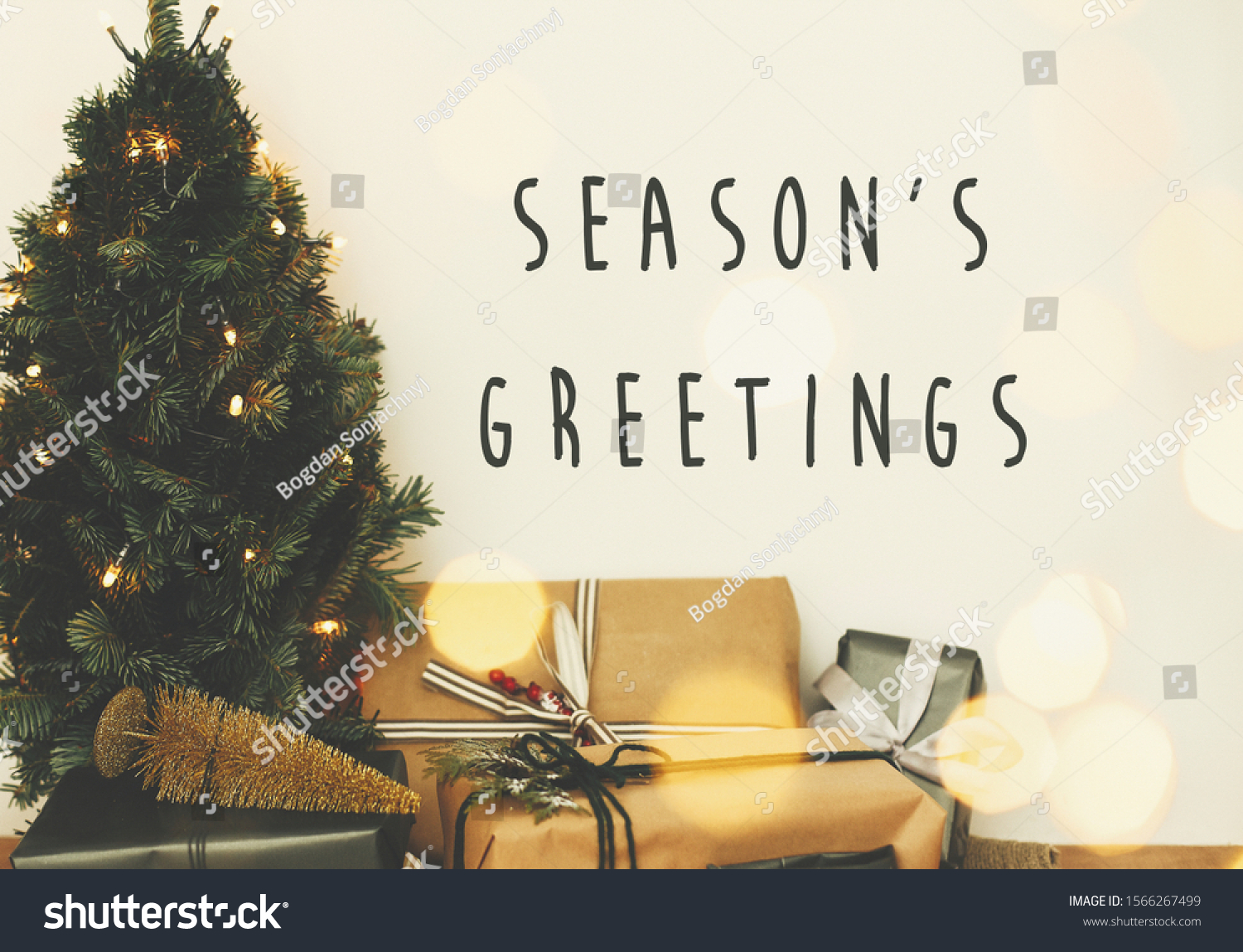 Season's greetings text sign on christmas tree in golden lights bokeh with festive stylish gifts in white room. Seasons greeting card #1566267499