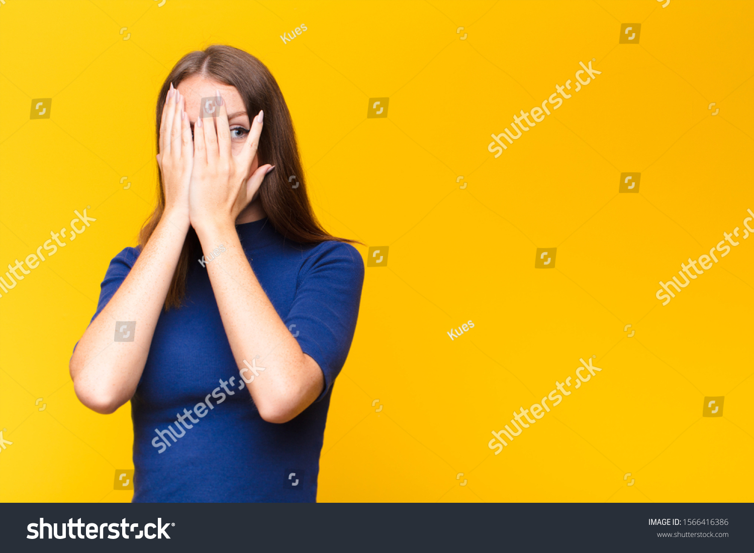 young red head woman covering face with hands, peeking between fingers with surprised expression and looking to the side against flat wall #1566416386