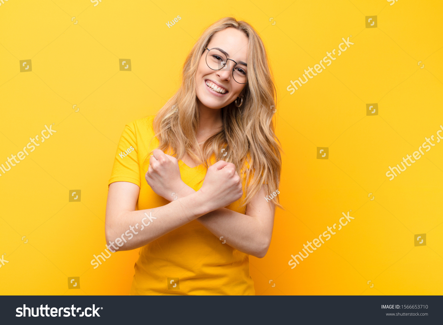 young pretty blonde woman smiling cheerfully and celebrating, with fists clenched and arms crossed, feeling happy and positive against flat color wall #1566653710