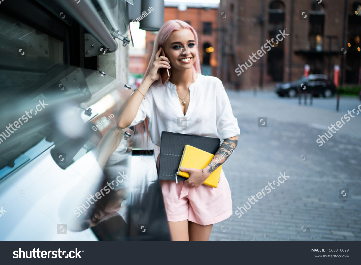 Young happy fashionable female in white shirt and rose shorts talking on mobile phone while walking in street with notebook and documents and looking at camera #1568816629