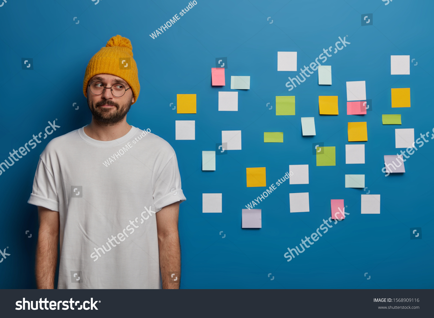 Indoor shot of serious contemplative man with beard, dressed casually, thinks about writing diploma, uses adhesive notes to write down information to remember. Male manager plans working day #1568909116