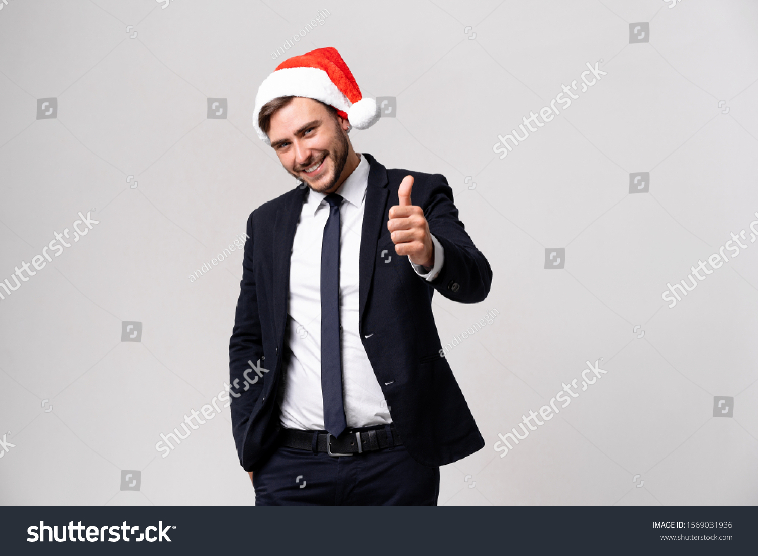 Young handsome caucasian guy in business suit and Santa hats on white background in studio smilie and showing thumbs up two hands. Close up portrait business person with Christmas mood Holiday banner #1569031936