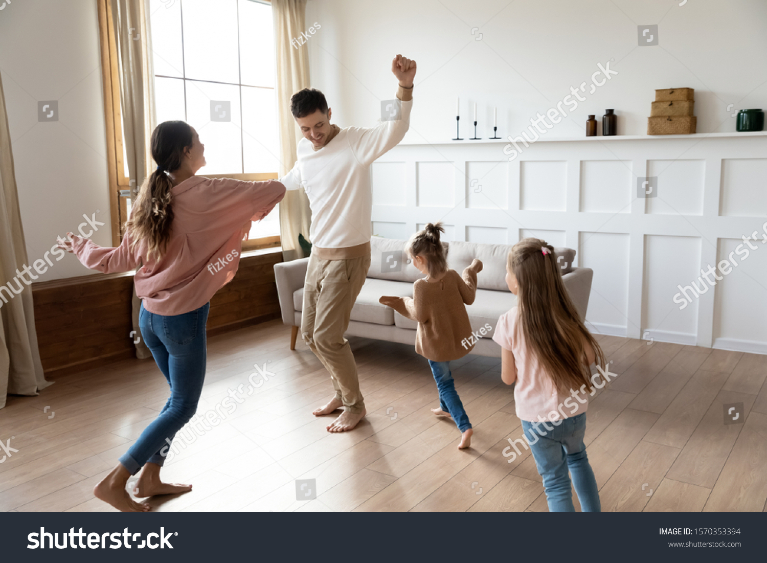 Funny active family of four young adult parents and cute small children daughters dancing together in living room interior, carefree little kids with mum dad having fun laughing enjoy leisure at home #1570353394