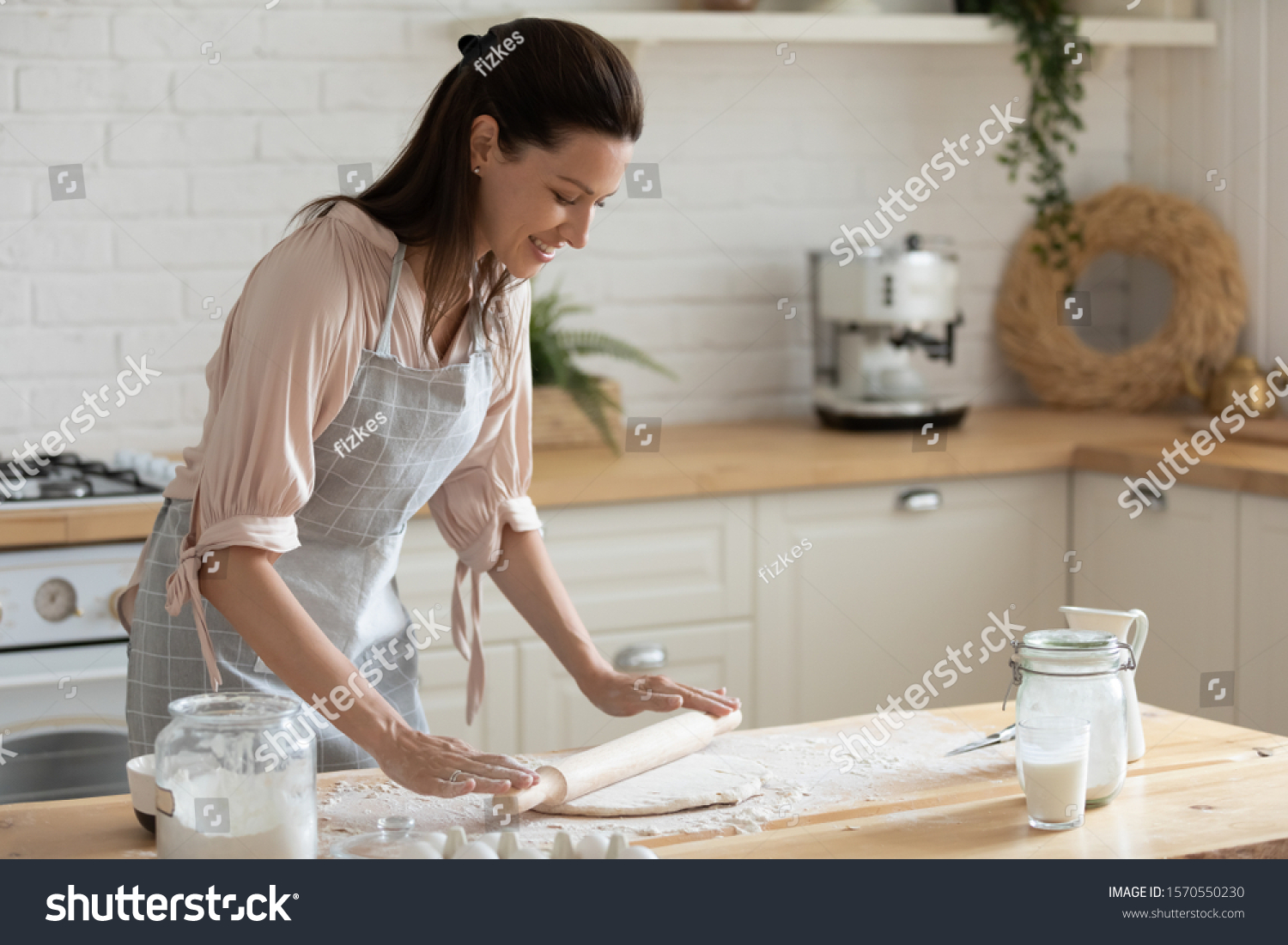 Happy attractive young adult woman lady housewife baker wear apron holding pin rolling dough on kitchen table baking pastry concept cooking cake biscuit doing bakery making homemade pizza at home #1570550230