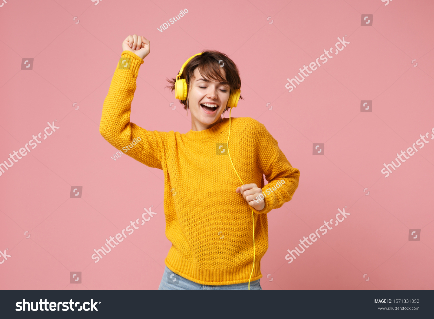 Cheerful young brunette woman girl in yellow sweater posing isolated on pastel pink wall background studio portait. People lifestyle concept. Mock up copy space. Listen music with headphones, dancing #1571331052