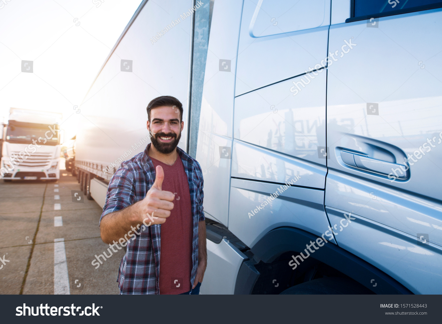 Portrait of young bearded man with thumbs up standing by his truck. Professional and positive truck driver standing by semi truck vehicle. Transportation services. #1571528443