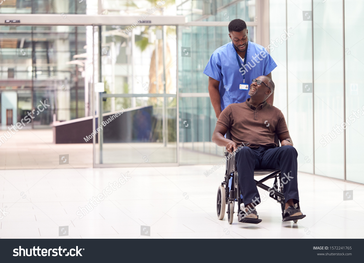 Male Nurse Wearing Scrubs Wheeling Patient In Wheelchair Through Lobby Of Modern Hospital Building #1572241765