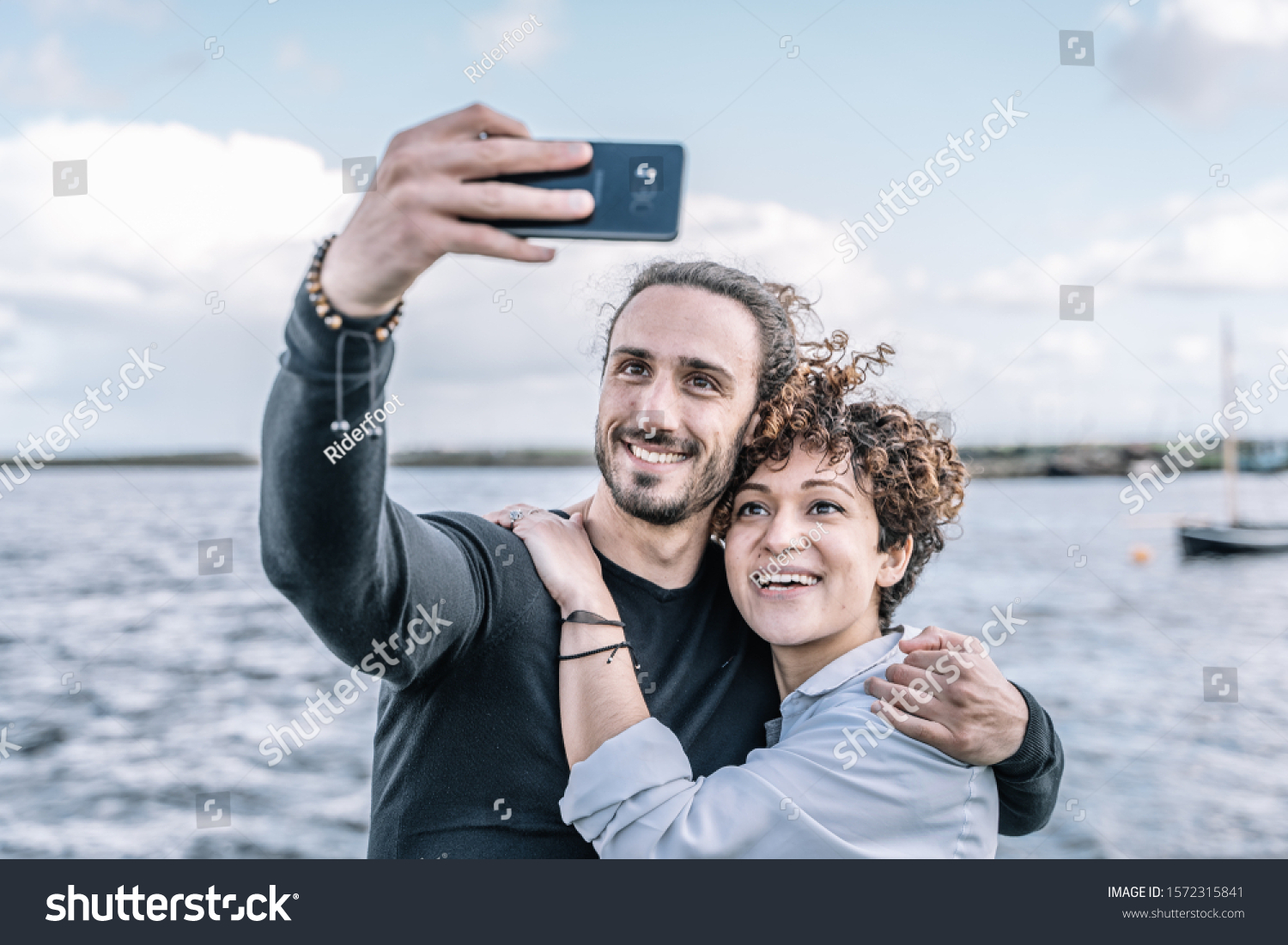 Stock photo of a young couple embraced by the shoulder making a selfie with the port and the sea out of focus background #1572315841