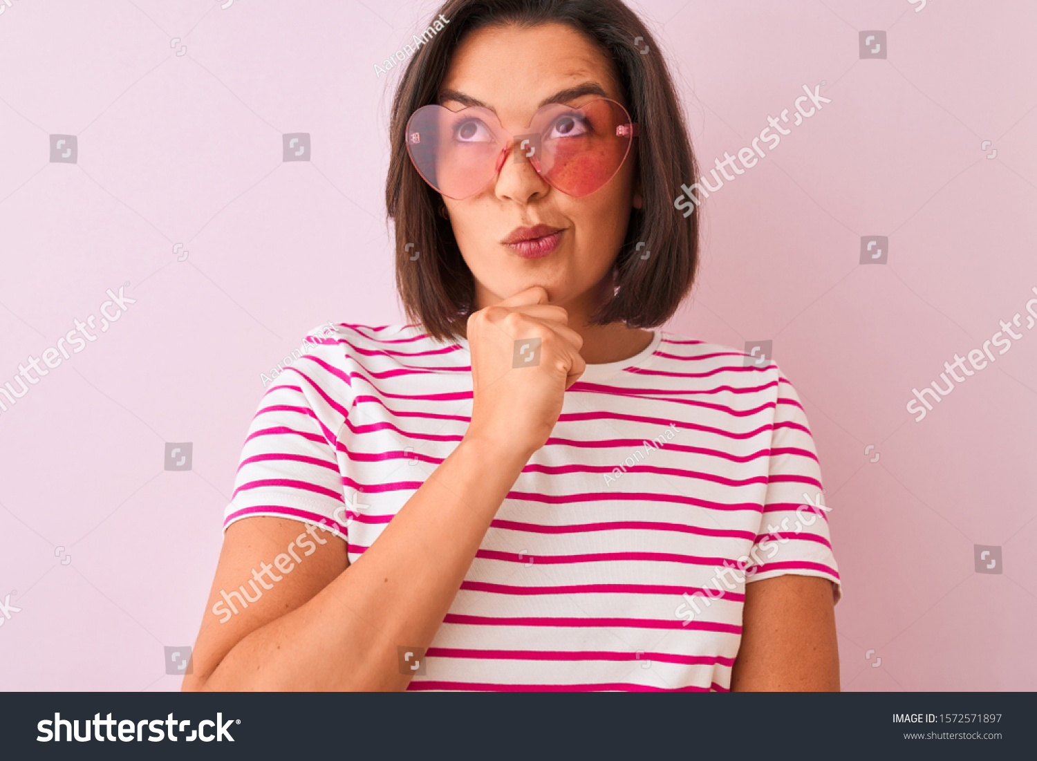 Young beautiful woman wearing fashion sunglasses with hearts over isolated pink background serious face thinking about question, very confused idea #1572571897