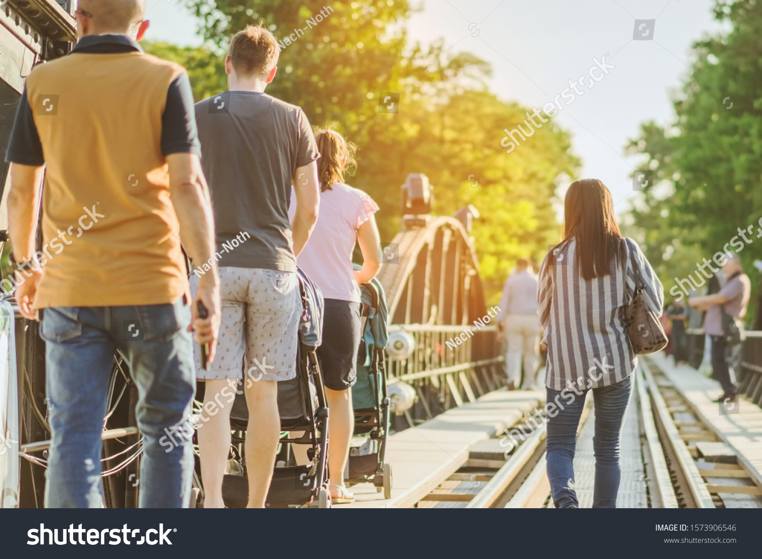 Dad and mum take their children on a stroller to walk and enjoy the beauty of The Bridge Over the River Kwai happily in the evening in Kanchanaburi, Thailand #1573906546