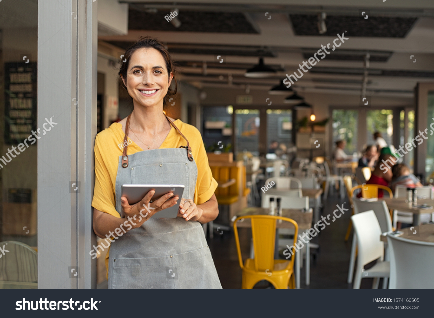 Portrait of happy woman standing at doorway of her store. Cheerful mature waitress waiting for clients at coffee shop. Successful small business owner in casual wearing grey apron standing at entrance #1574160505