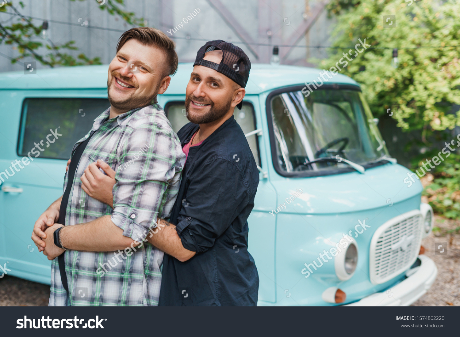 Portrait of a gay couple in front of an old-fashioned car van. Boyfriends hug and smile to the camera. #1574862220