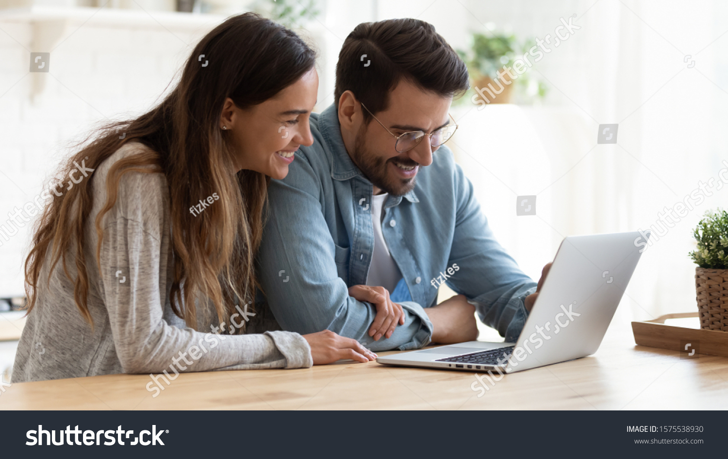 Happy young family couple bonding laughing using laptop looking at screen sit at table, smiling husband and wife buying choosing goods online doing internet shopping order with delivery at home #1575538930
