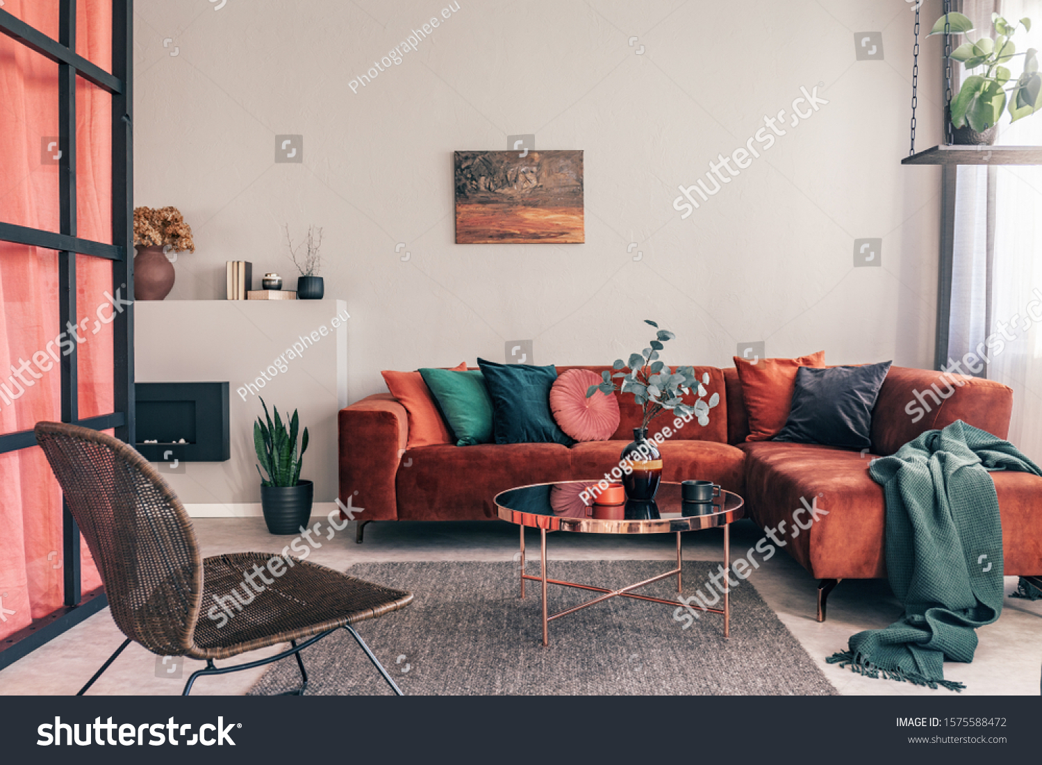 Real photo of a comfy living room interior with a round table on gray rug, wicker armchair and red corner sofa #1575588472