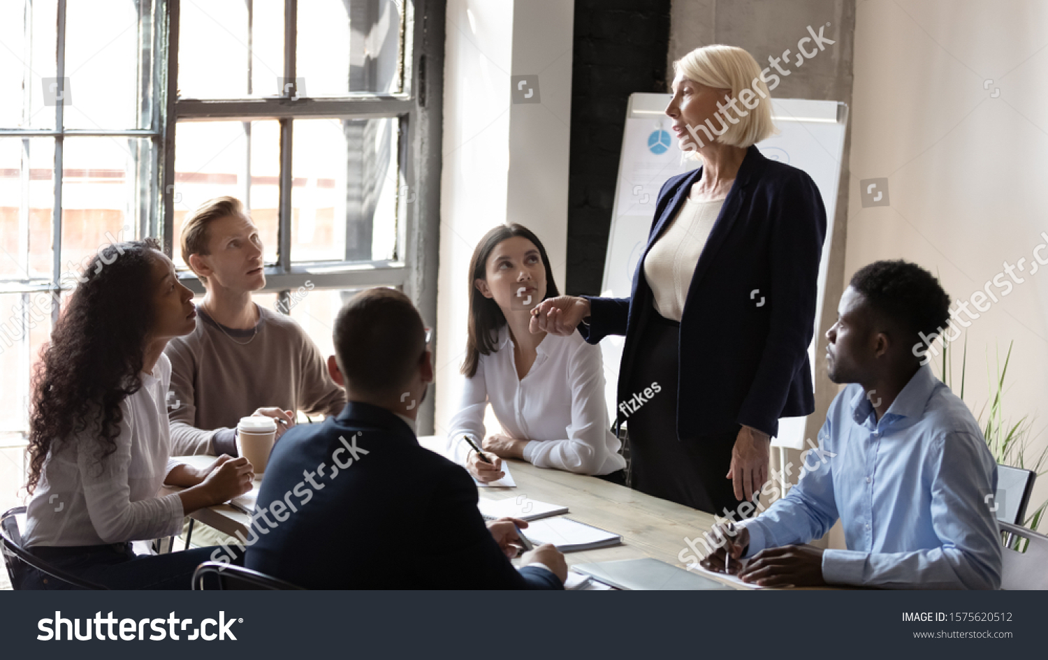 Middle aged professional coach educating focused mixed race managers at office. Concentrated young multiracial coworkers listening to older speaker, presenting project ideas at brainstorming meeting. #1575620512
