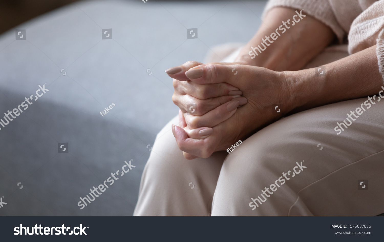 Elderly woman seated on couch holding hands together folded on lap feeling lonely and abandoned close up image. Solitude or loneliness, person recollect memories, waiting, life in nursing home concept #1575687886