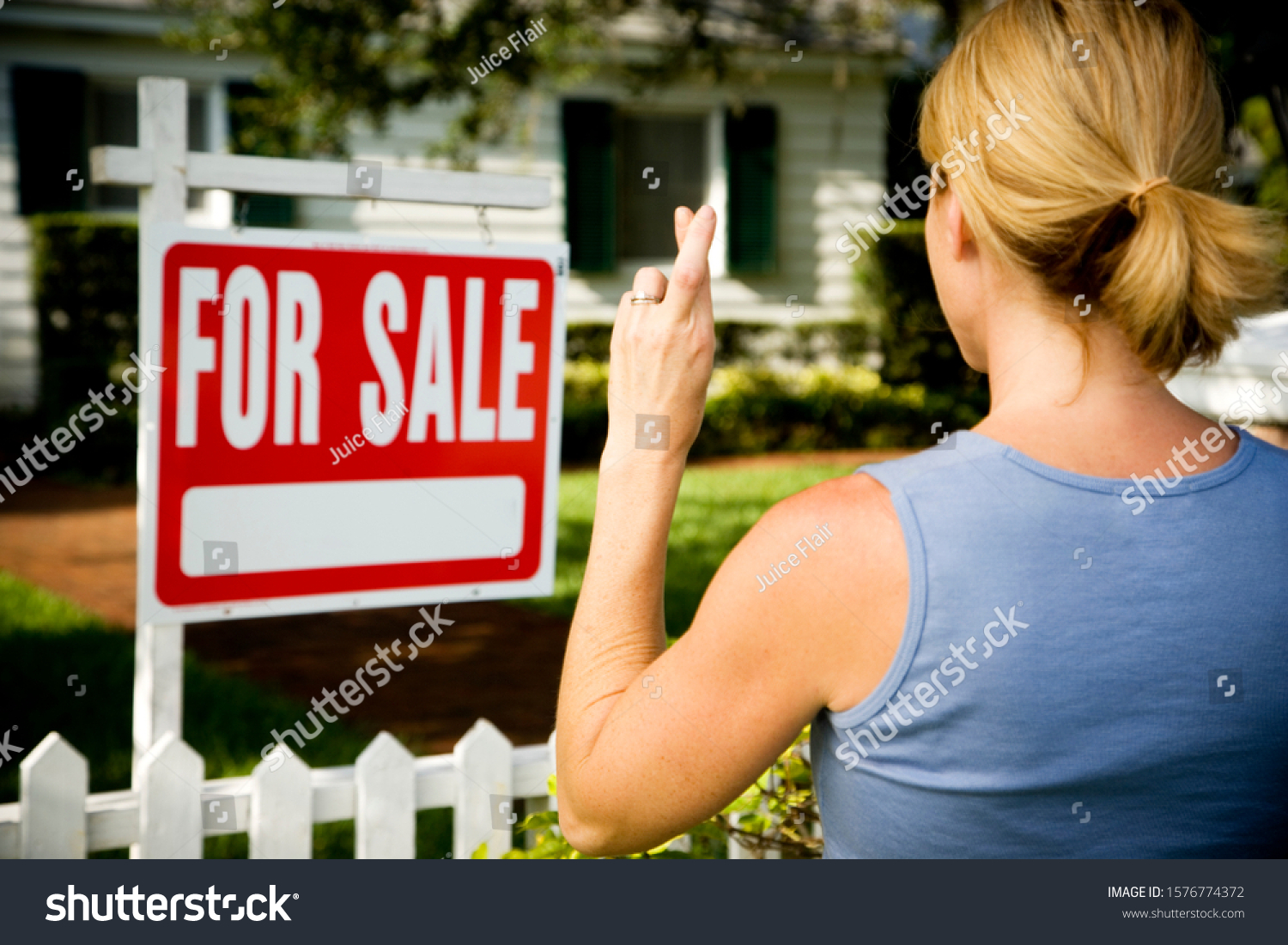 Woman standing by a for sale sign outside a family house, fingers crossed #1576774372