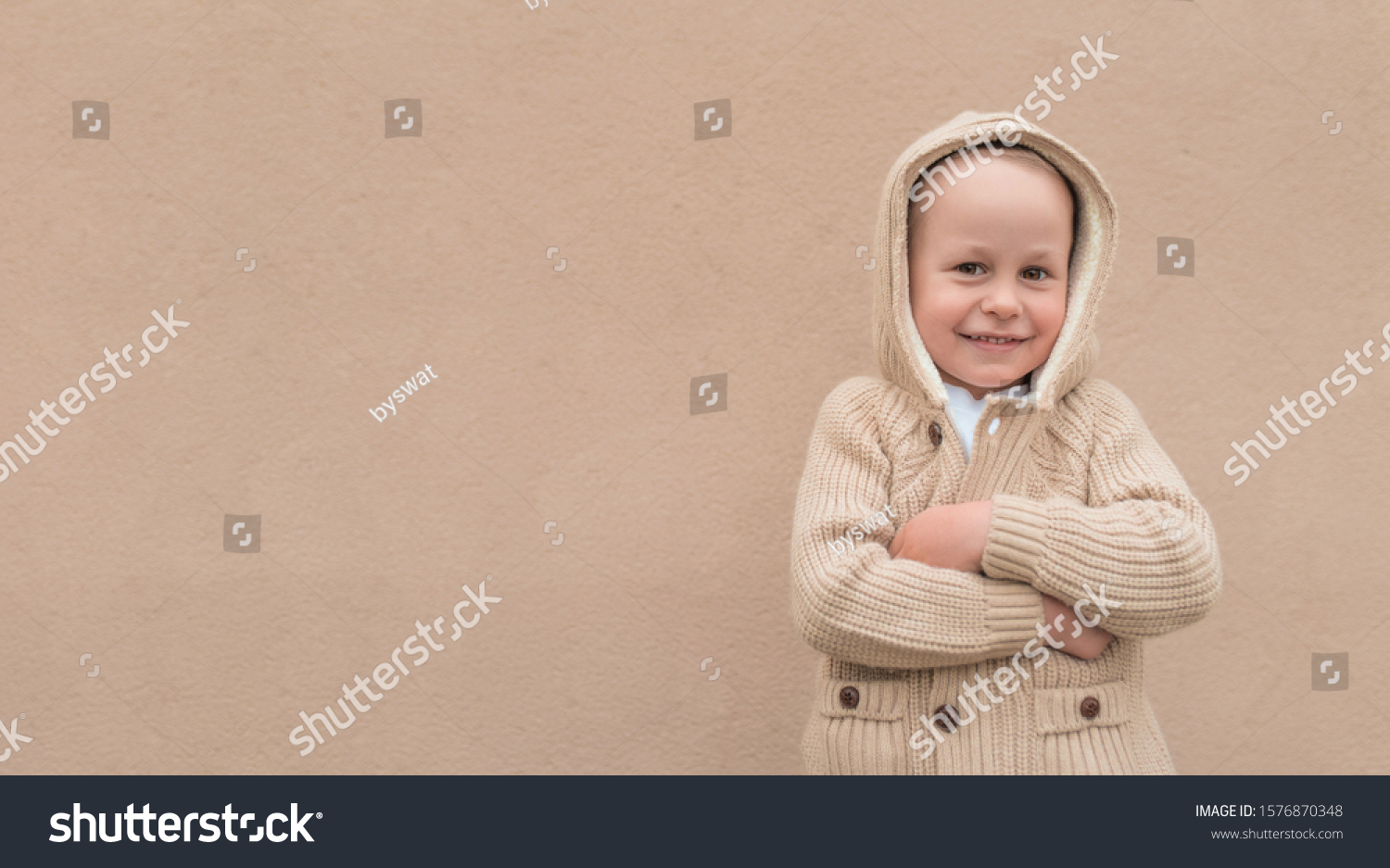 Little boy 3-5 years old, autumn beige wall, in warm casual clothes, sweater with hood, happy smiling, free space for copy text. Christmas mood of happiness fun and relaxation. Holiday concept #1576870348