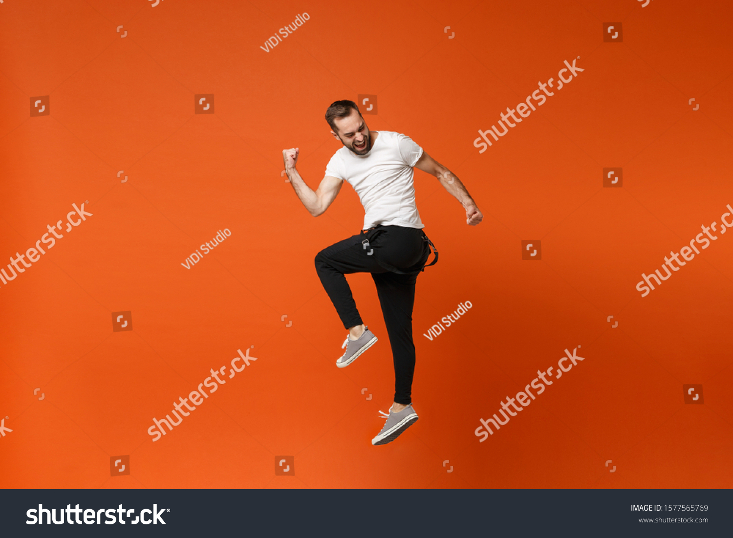 Cheerful young man in casual white t-shirt posing isolated on bright orange wall background studio portrait. People lifestyle concept. Mock up copy space. Having fun, jumping, doing winner gesture #1577565769
