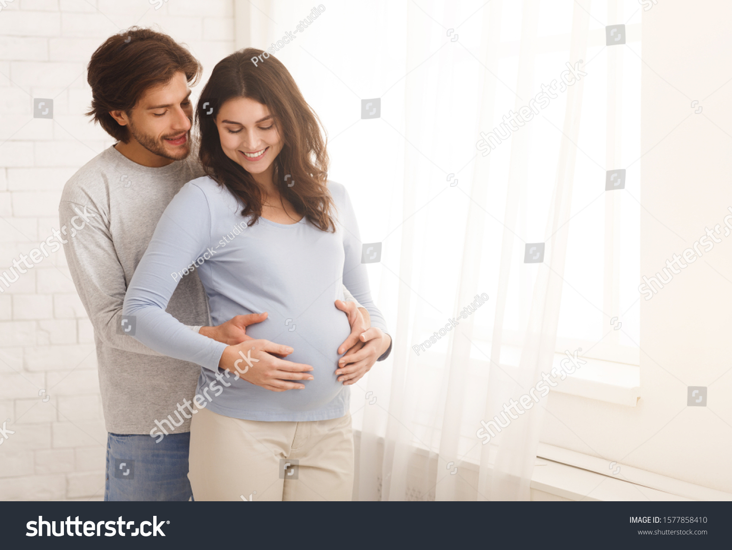 Loving man hugging his pregnant wife from behind standing near window at home, copy space #1577858410