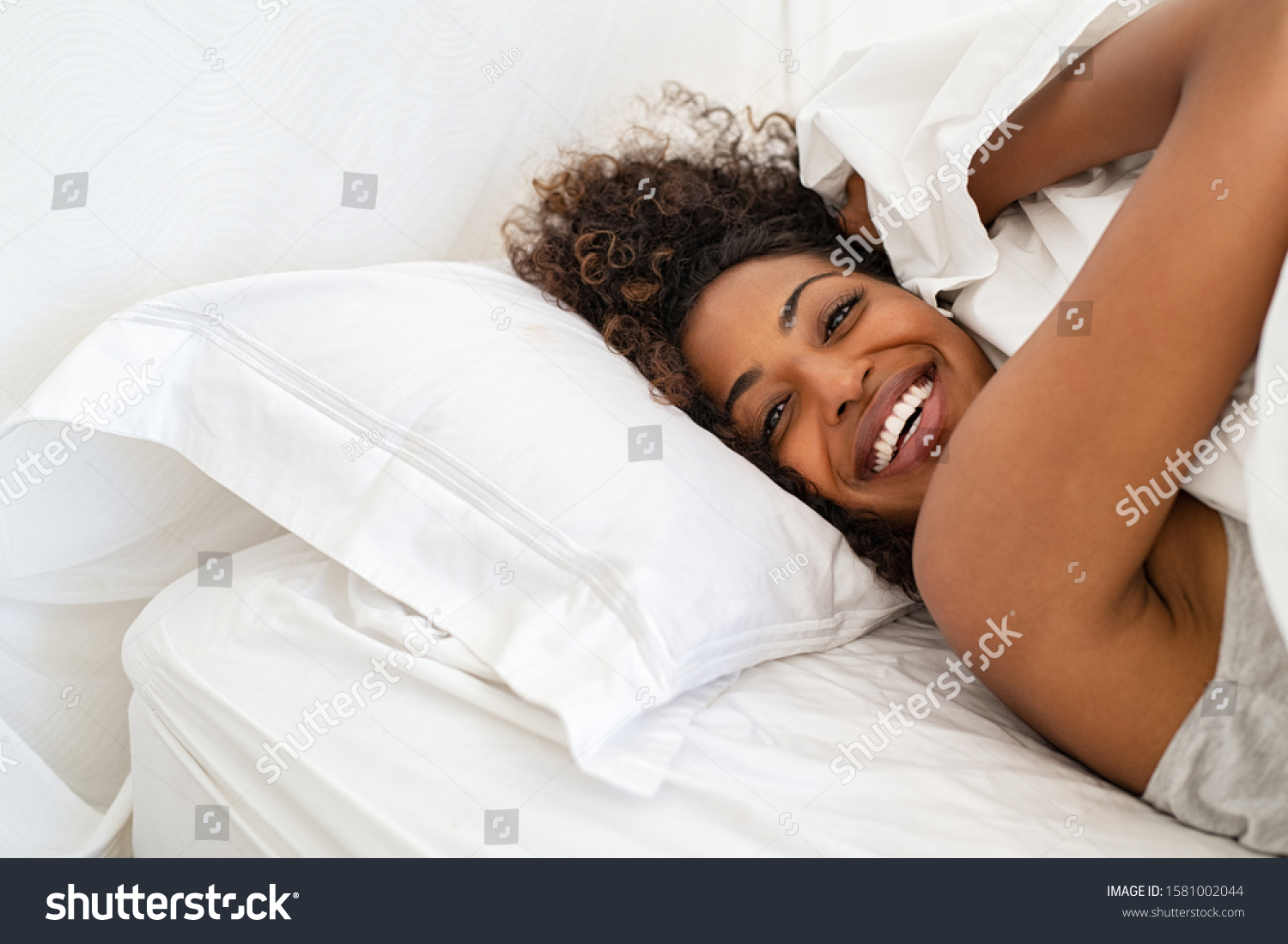 Cheerful young woman lying on bed playing with blanket and looking at camera. African girl feeling fresh after nap on bed with copy space. Laughing woman having fun while embracing pillow and blanket. #1581002044