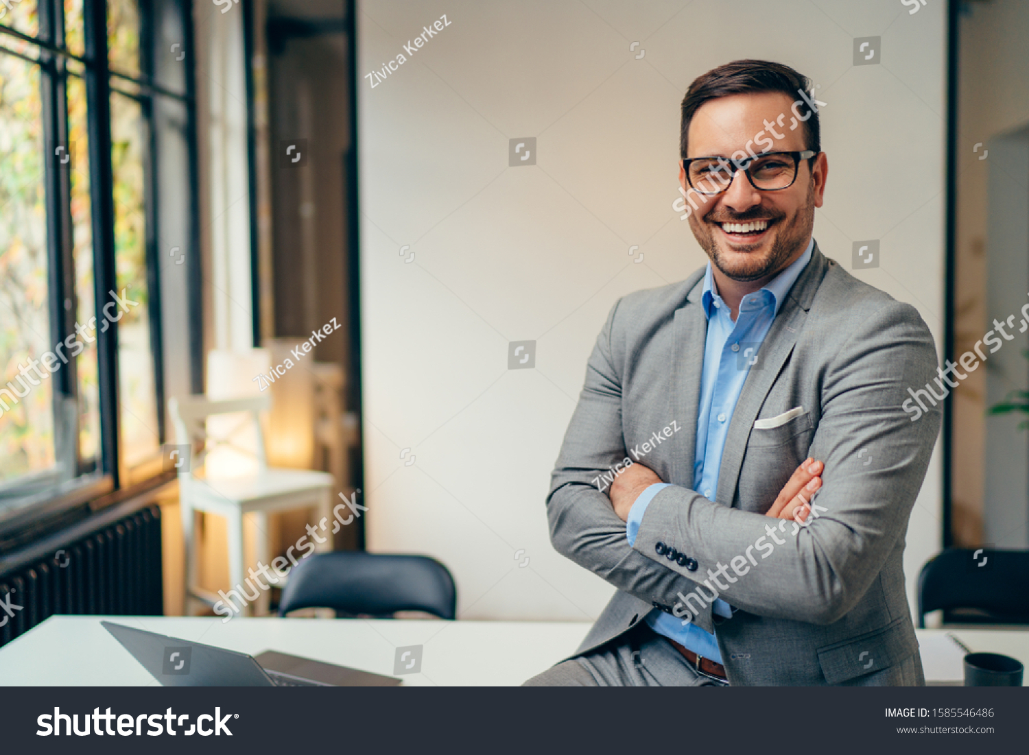 Portrait of young happy businessman wearing grey suit and blue shirt standing in his office and smiling with arms crossed #1585546486