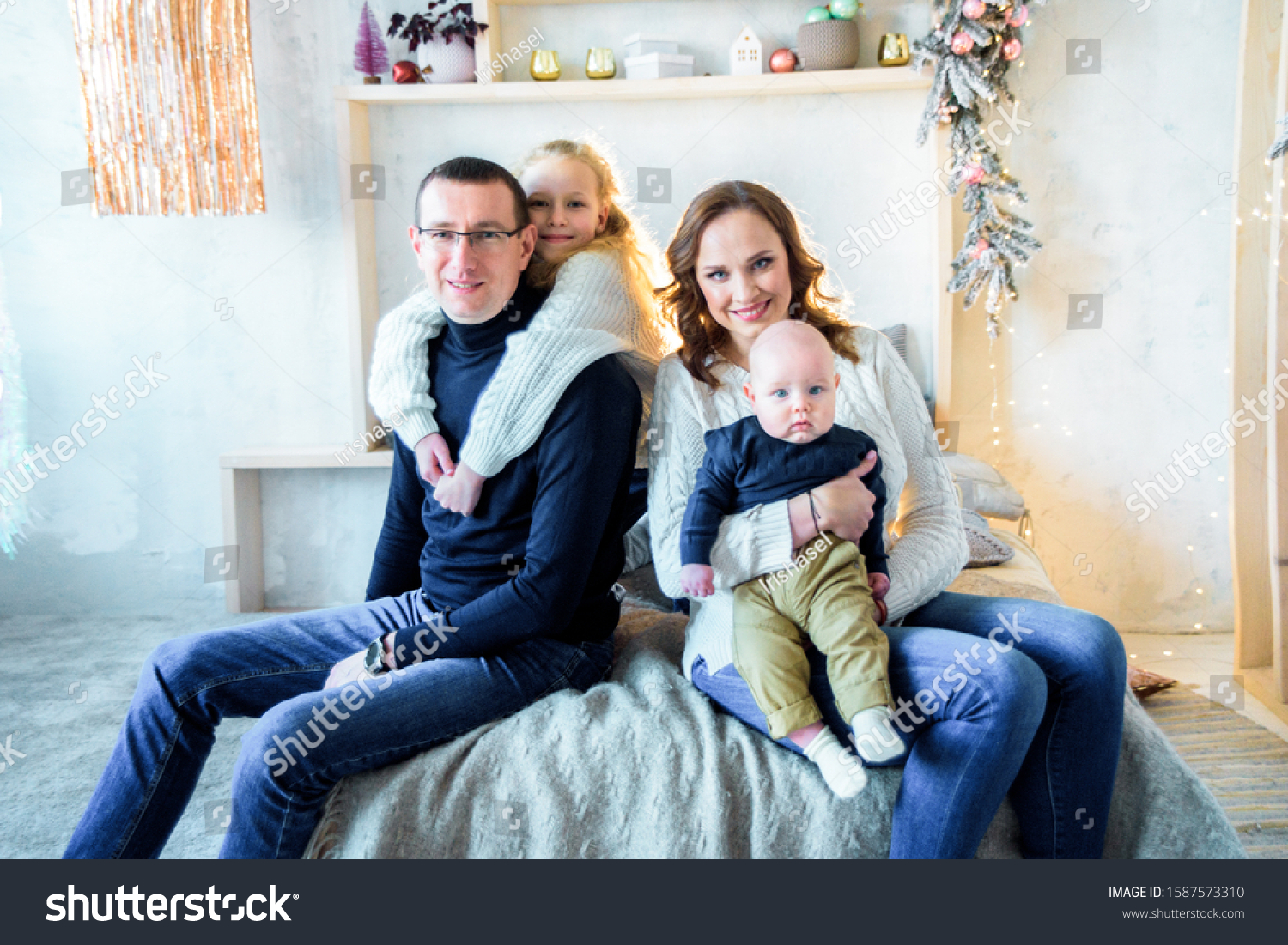 Happy mom and dad, sister and little brother on the bed in a large bright bedroom. Christmas decor and lights. Big happy family. The concept of parental love and parenting. #1587573310