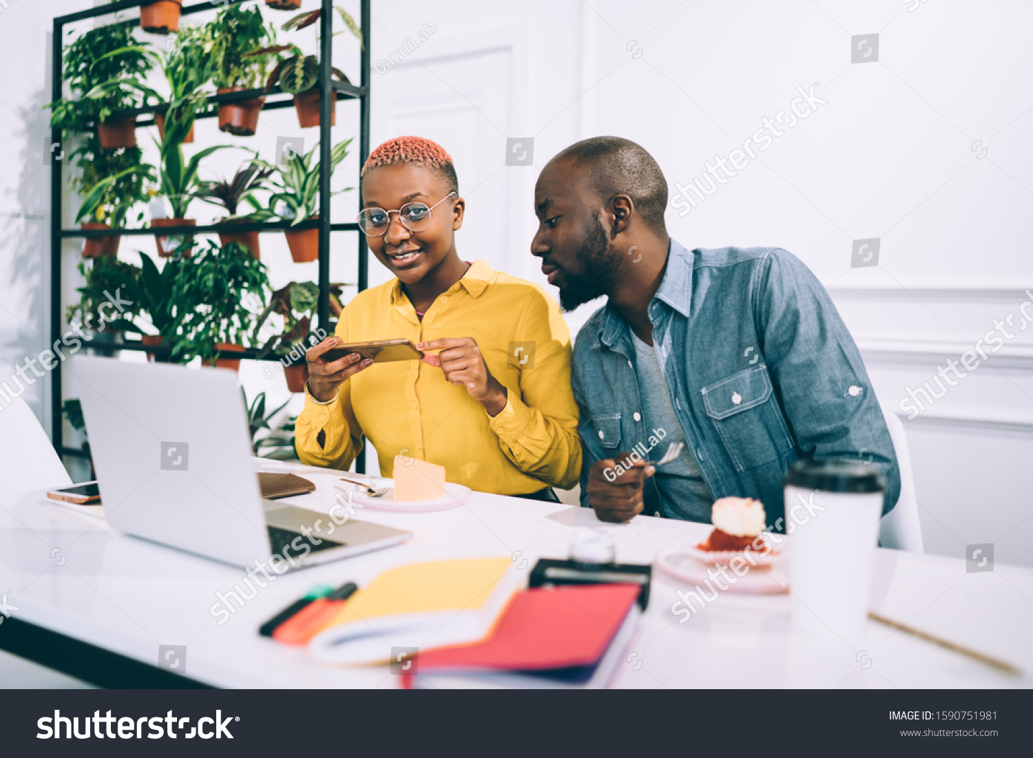 Young African American woman taking photo of piece of cake with smartphone for sharing in social media while working with male colleague in cafeteria #1590751981