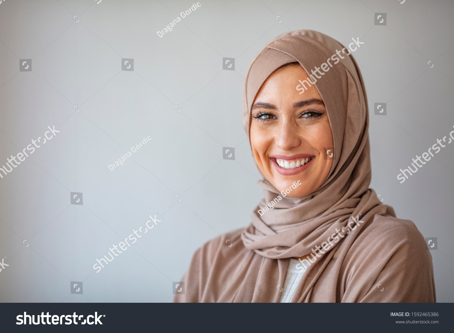Woman in traditional Muslim clothing, smiling. Beautiful woman headshot looking at camera and wearing a hijab. Arabian woman with happy smile. Strict formal outfit and elegant appearance. Islamic #1592465386